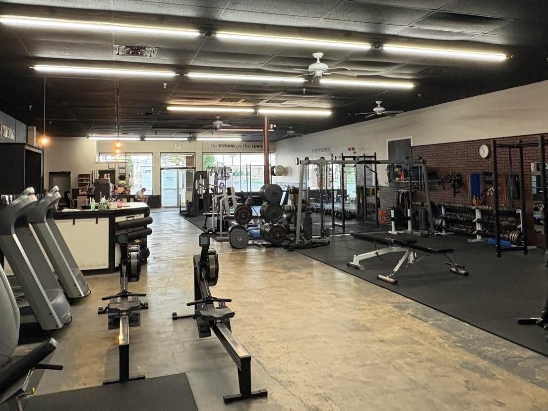 Rowers and treadmills facing the front desk, with racks and dumbbells along the wall